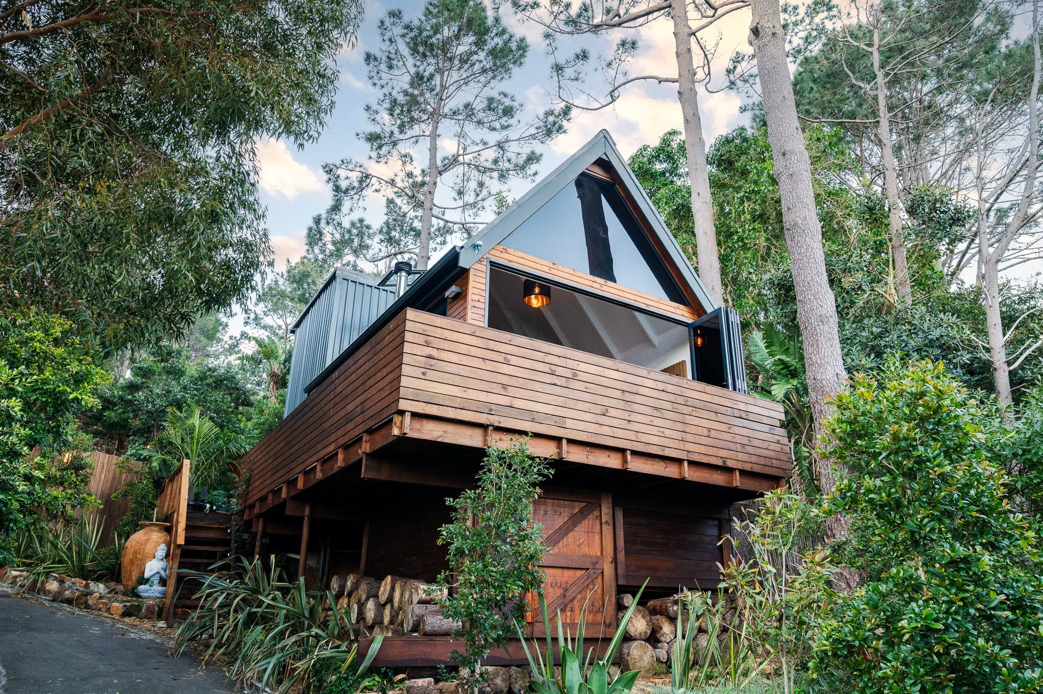 The A-frame cabin on stilts at dusk, its glazed gable glowing above the log store