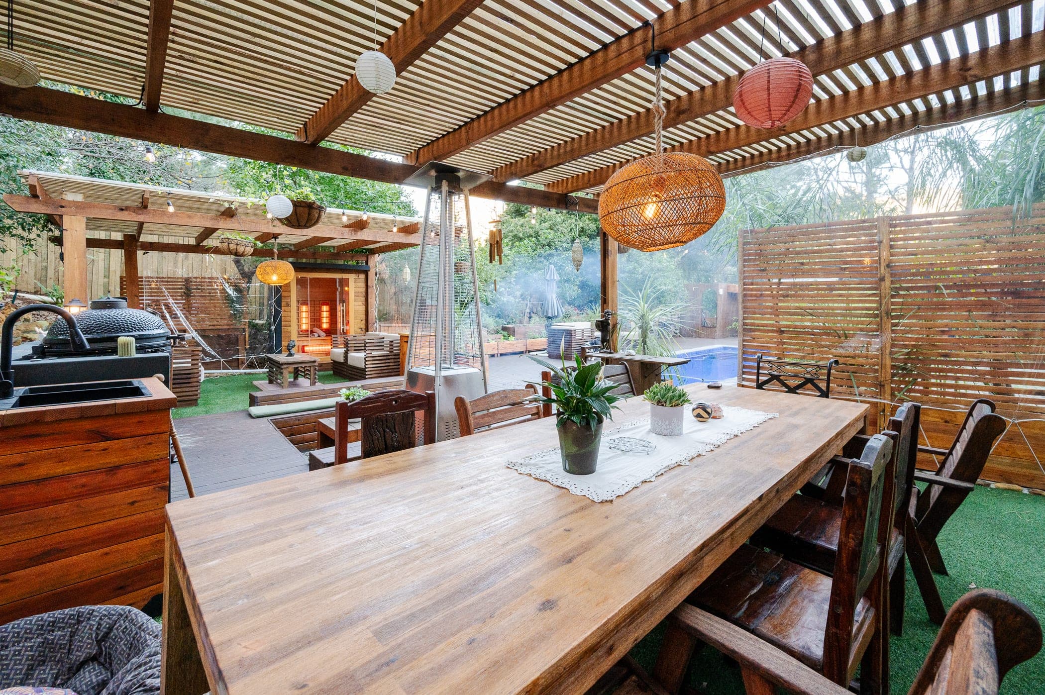 The long dining table under the slatted pergola, lanterns overhead